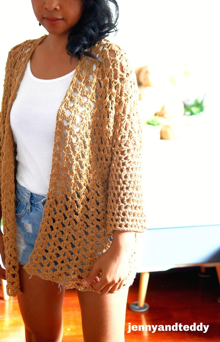  Dark haired woman wearing a yellow loosely weaved crochet cardigan with long sleeves over a white vest and denim shorts.