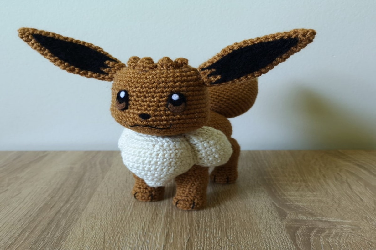 Small stuffed crochet Eevee with a white body, a brown leg, head and ears sticking out. Eevee is standing on a wooden table.
