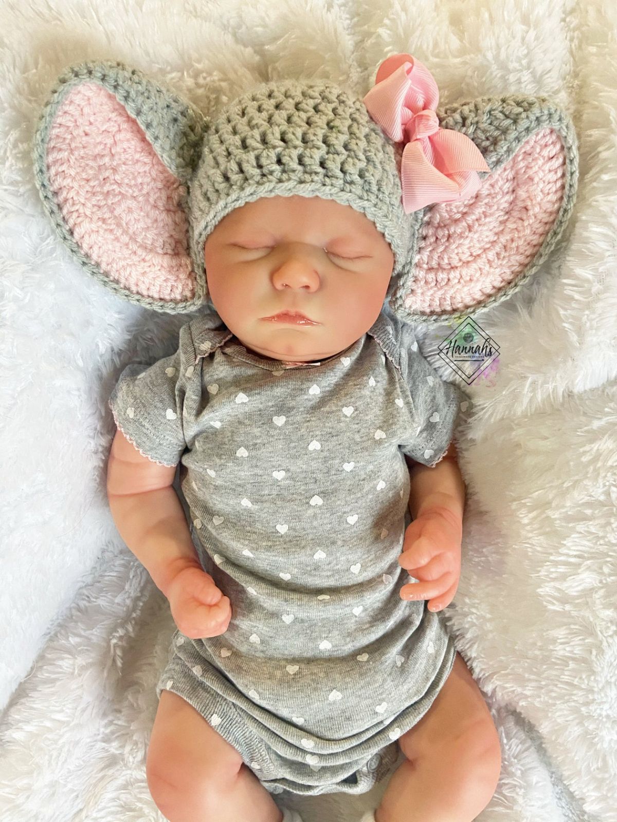 Newborn baby asleep wearing a gray crochet beanie with large gray and pink elephant ears on either side and a pink bow on the right ear.