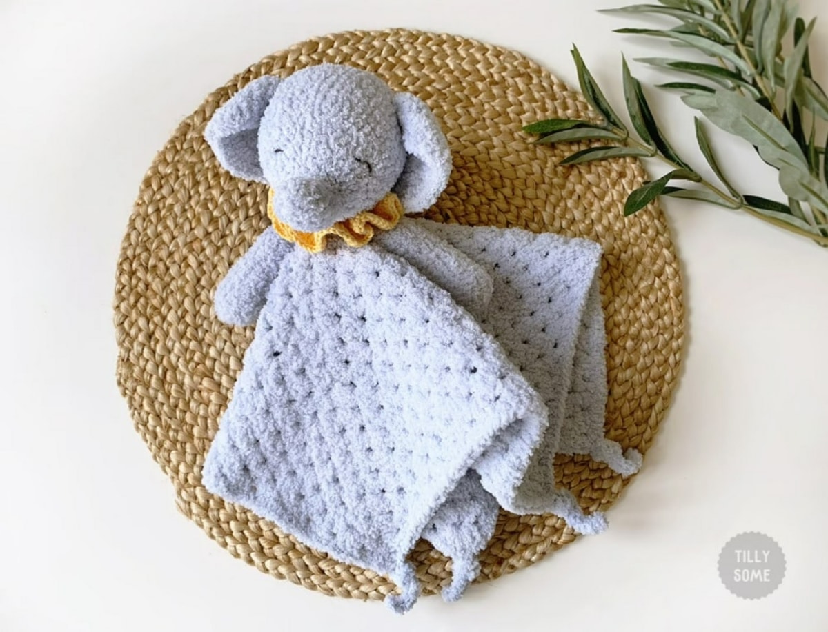 Small gray crochet elephant head and arms attached to a small baby blanket lying on a brown placemat.