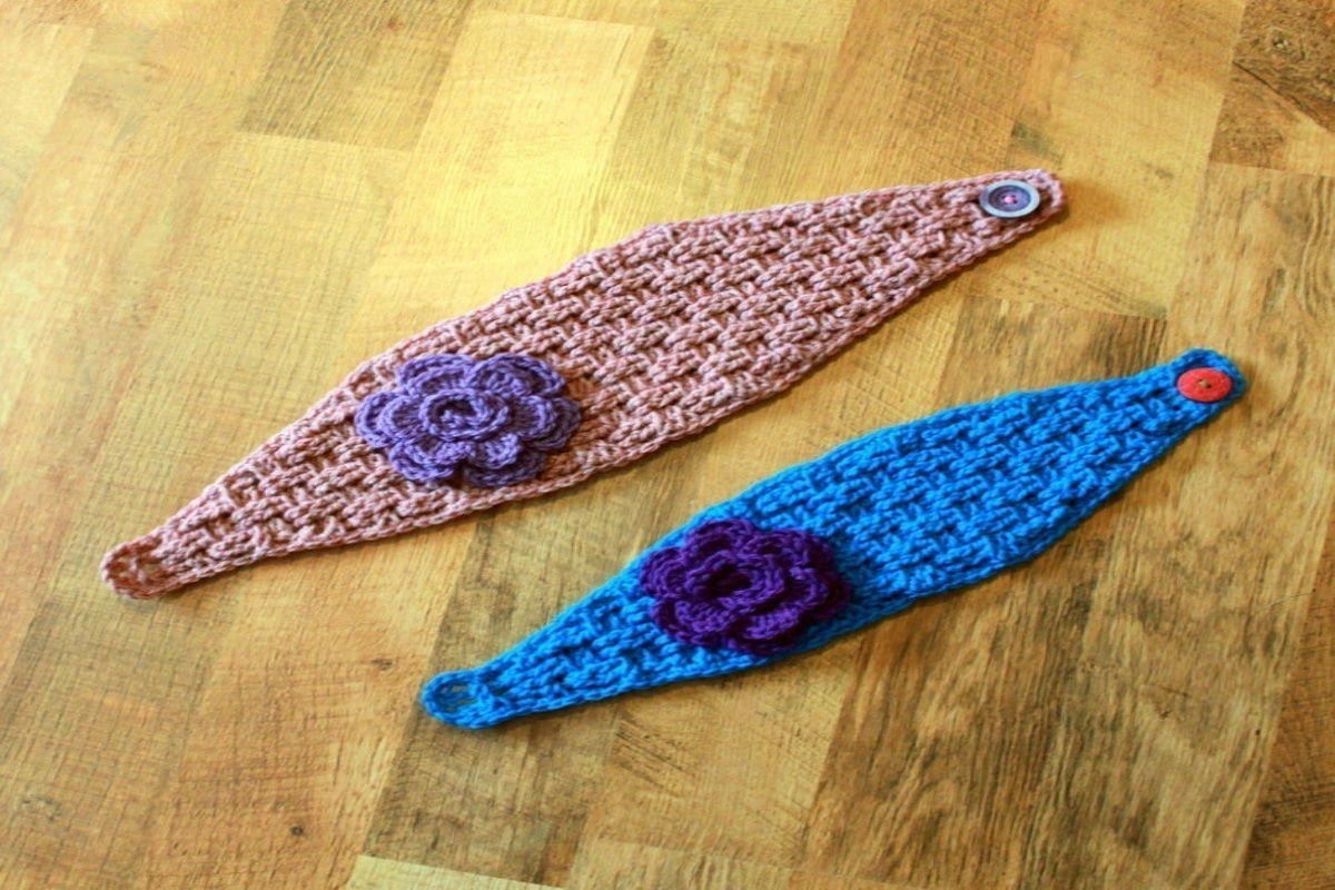 A blue crochet headwrap with a large purple flower on the side next to a beige headwrap with a purple flower on its side.