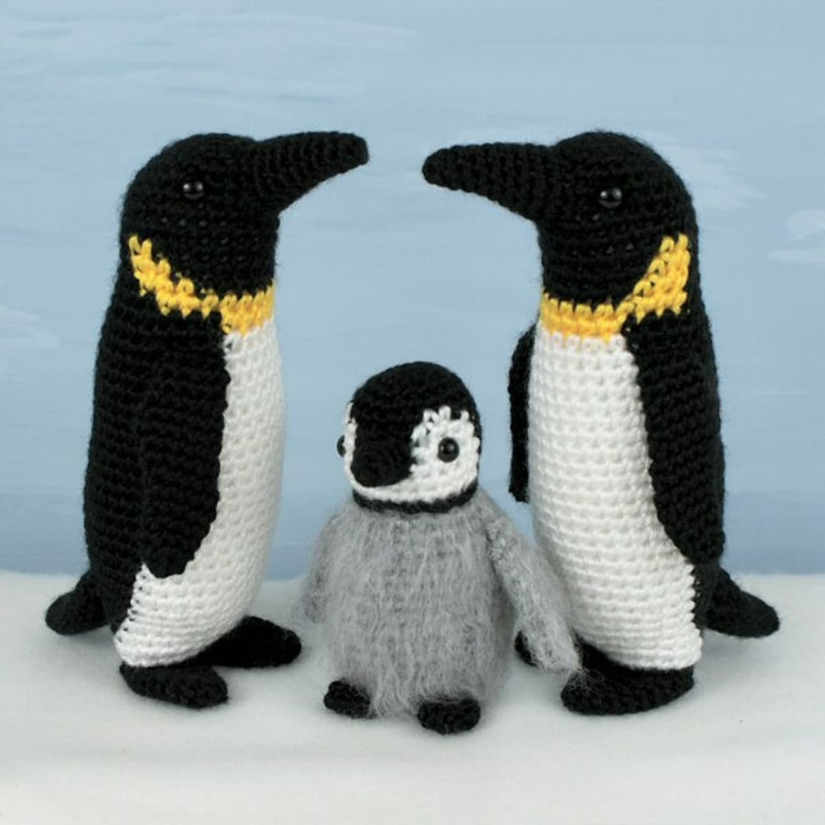 Two black and white crochet Emperor Penguins standing facing each other with a gold band around their neck and a baby penguin in the middle with a gray body.