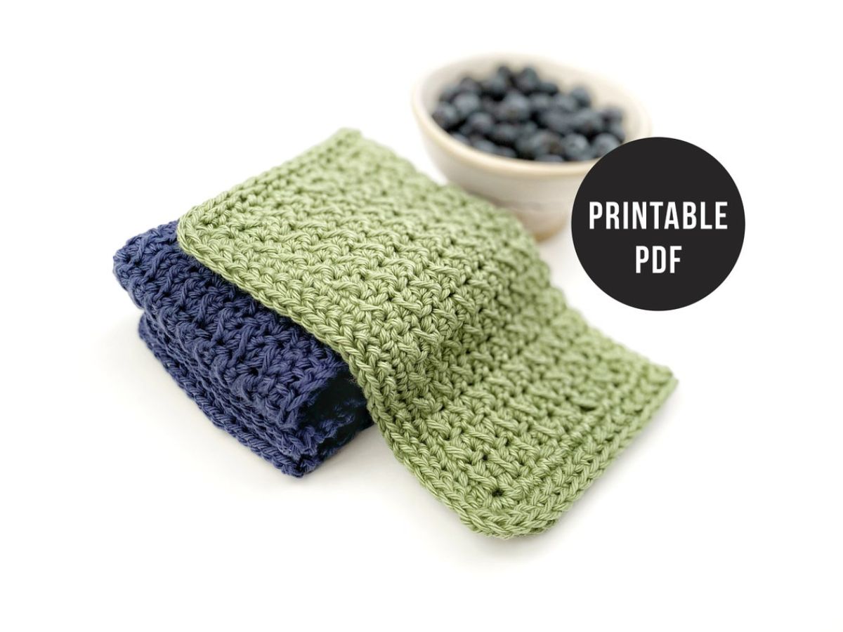A green crochet dishcloth spread over a folded navy blue dishcloth next to a small white bowl of pebbles.