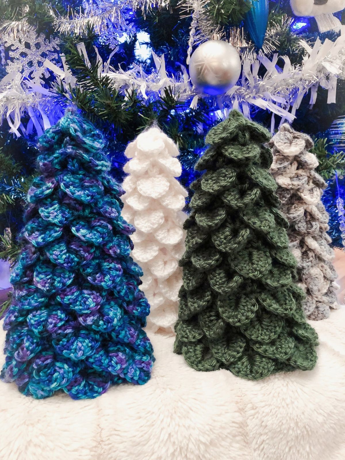 Four evergreen style crochet Christmas trees in green, white, gray, and purple and blue standing on a cream blanket.