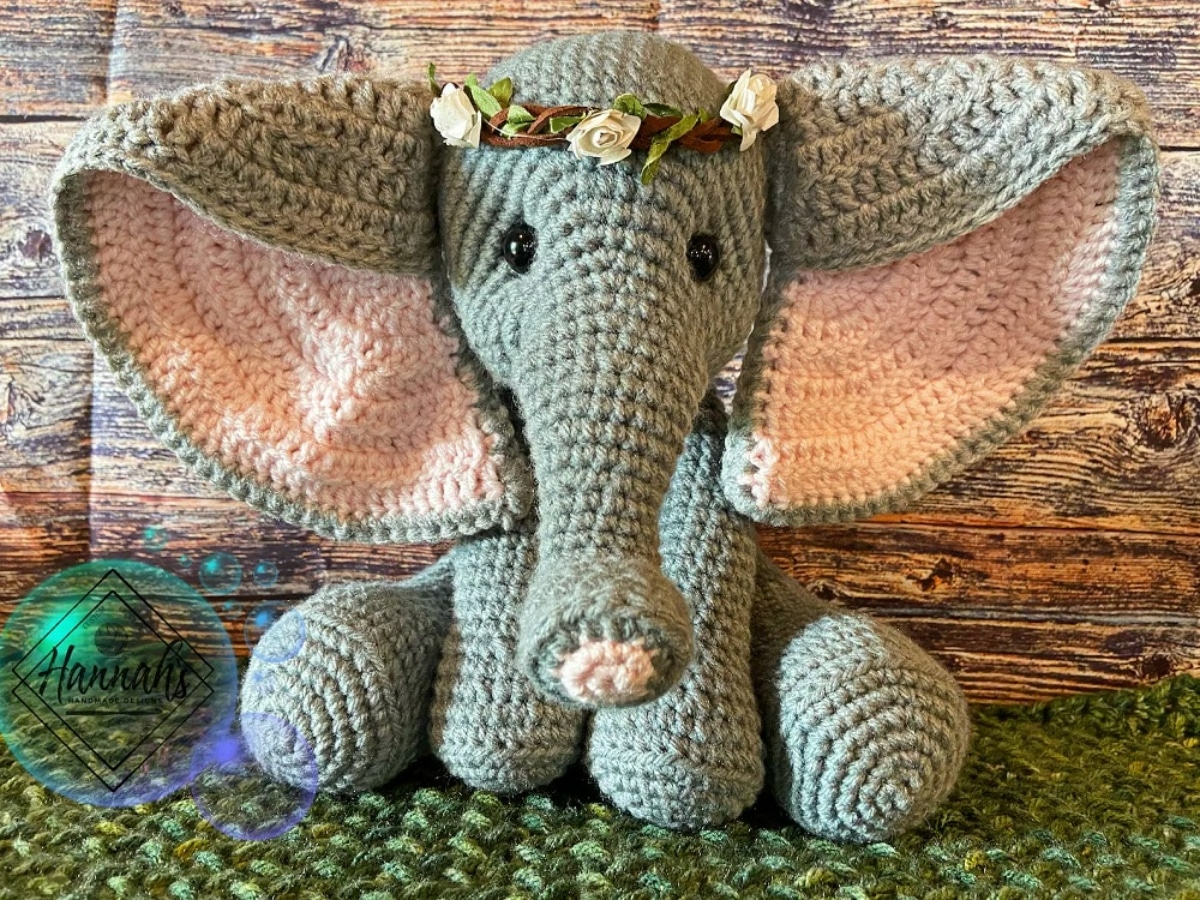 A large gray crochet Dumbo with large pink ears and a small brown and pink flower crown on its head sitting on a green blanket.