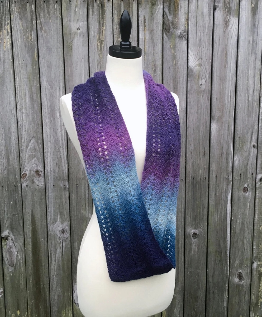 Purple, gray and blue ombre style crochet infinity scarf on a mannequin next to a gray wooden fence.