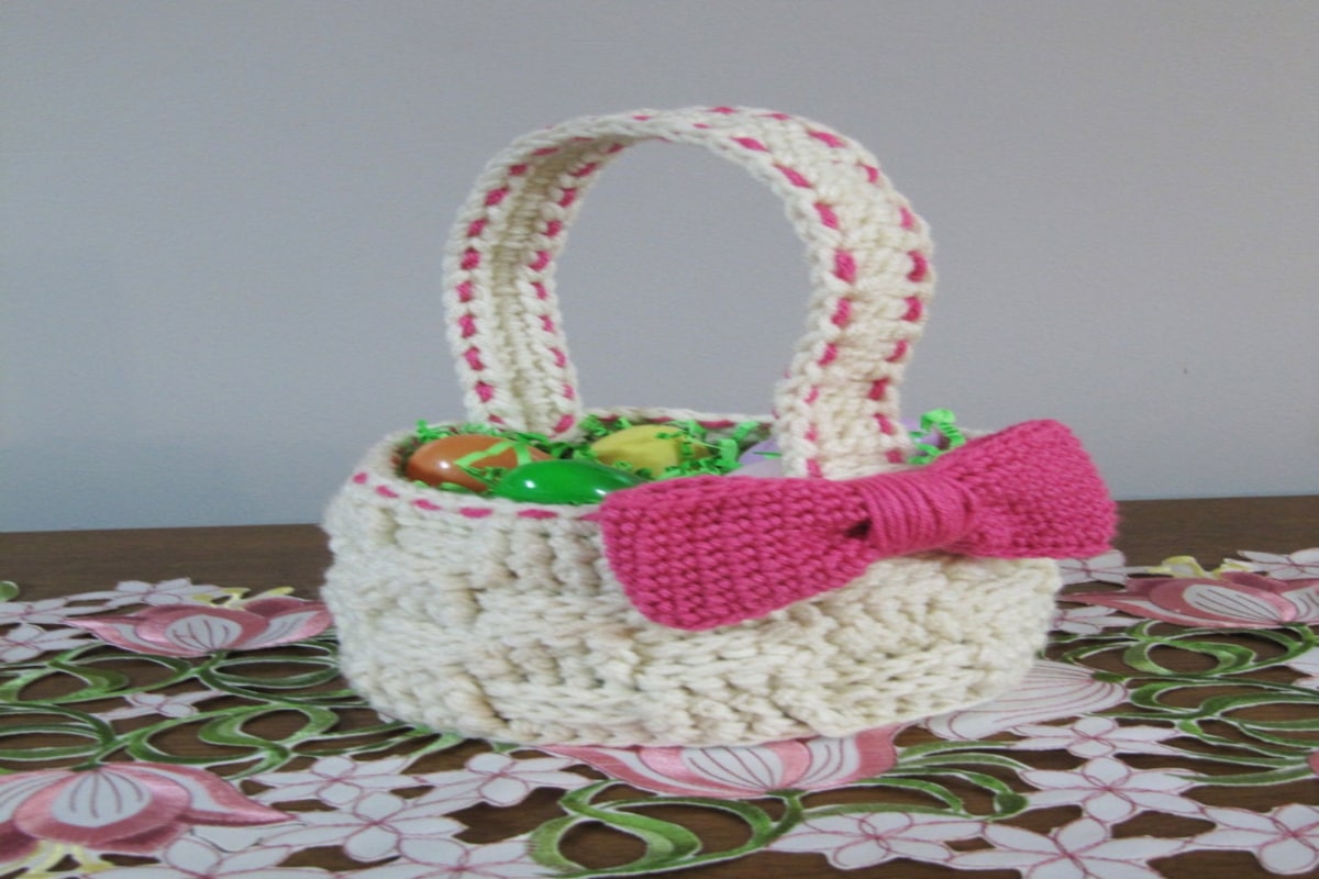 A short white crochet basket with a large pink bow on the right side and a thick handle filled with crochet Easter eggs.