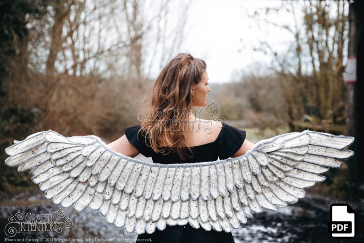 Brunette woman standing outside with a light gray feather style crochet shawl that resembles an angel's wings. 