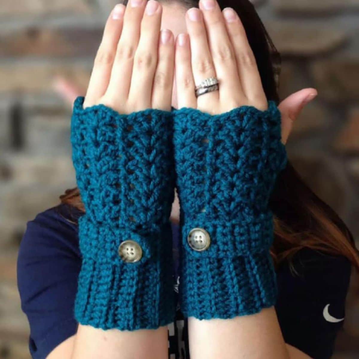 Dark blue crochet fingerless gloves with a wavy top and buttons around the wrist to secure worn by a woman with hands over her face. 