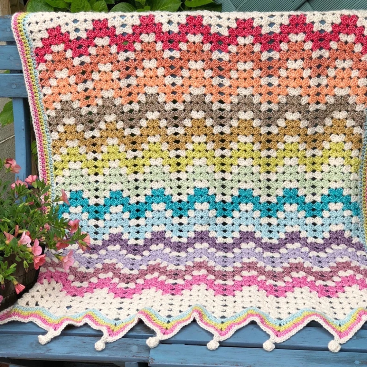 Rainbow colored crochet blanket using a granny ripple stitch with small white bobbles along the bottom draped over a blue bench.