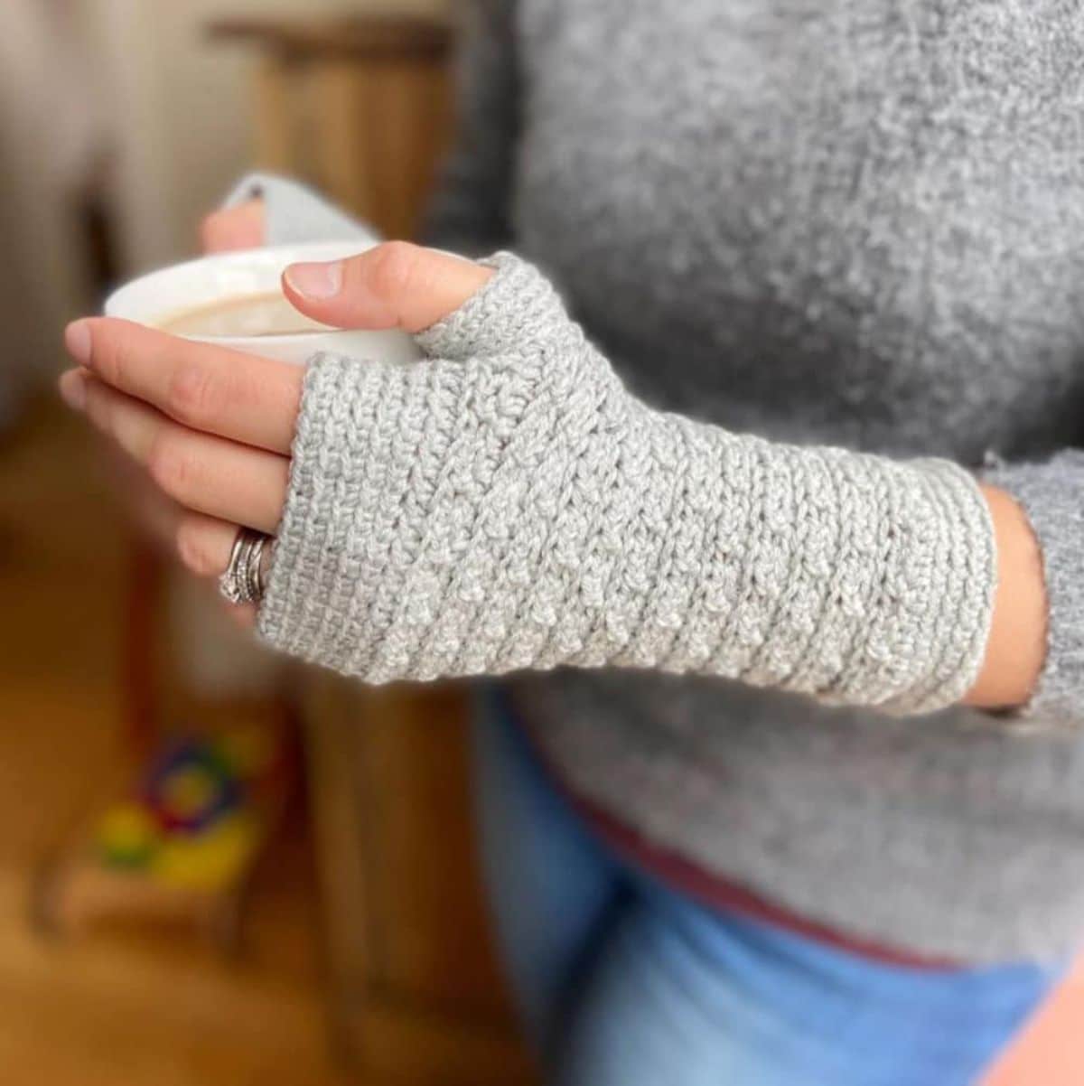 A woman in a gray jumper holding a white cup of coffee wearing gray crochet fingerless gloves.