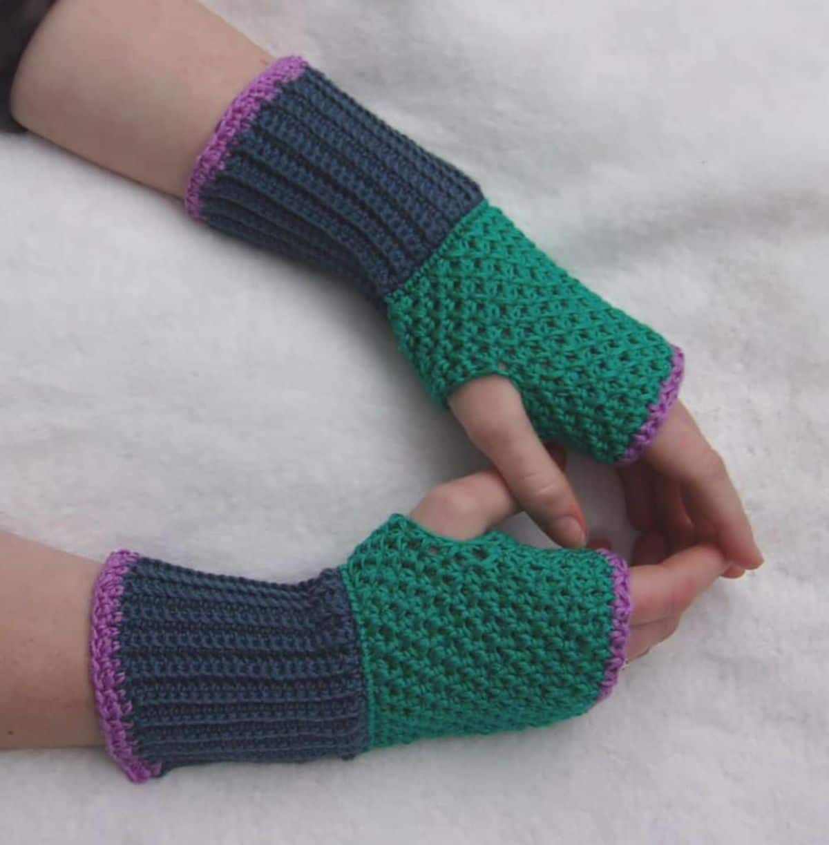 Purple and green fingerless gloves with the colors in large blocks and worn by hands on a white background.