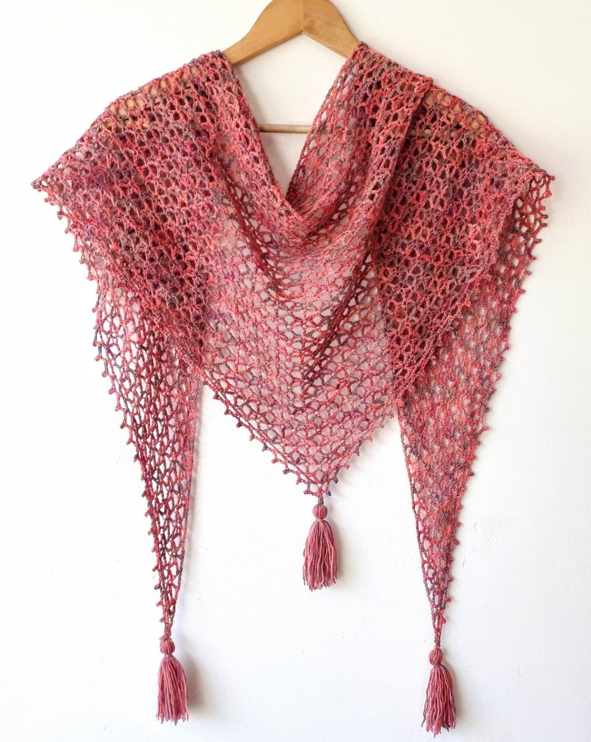 Pink crochet fishnet style lace shawl with a pink tassel hanging from each corner of the shawl draped over a wooden hanger.