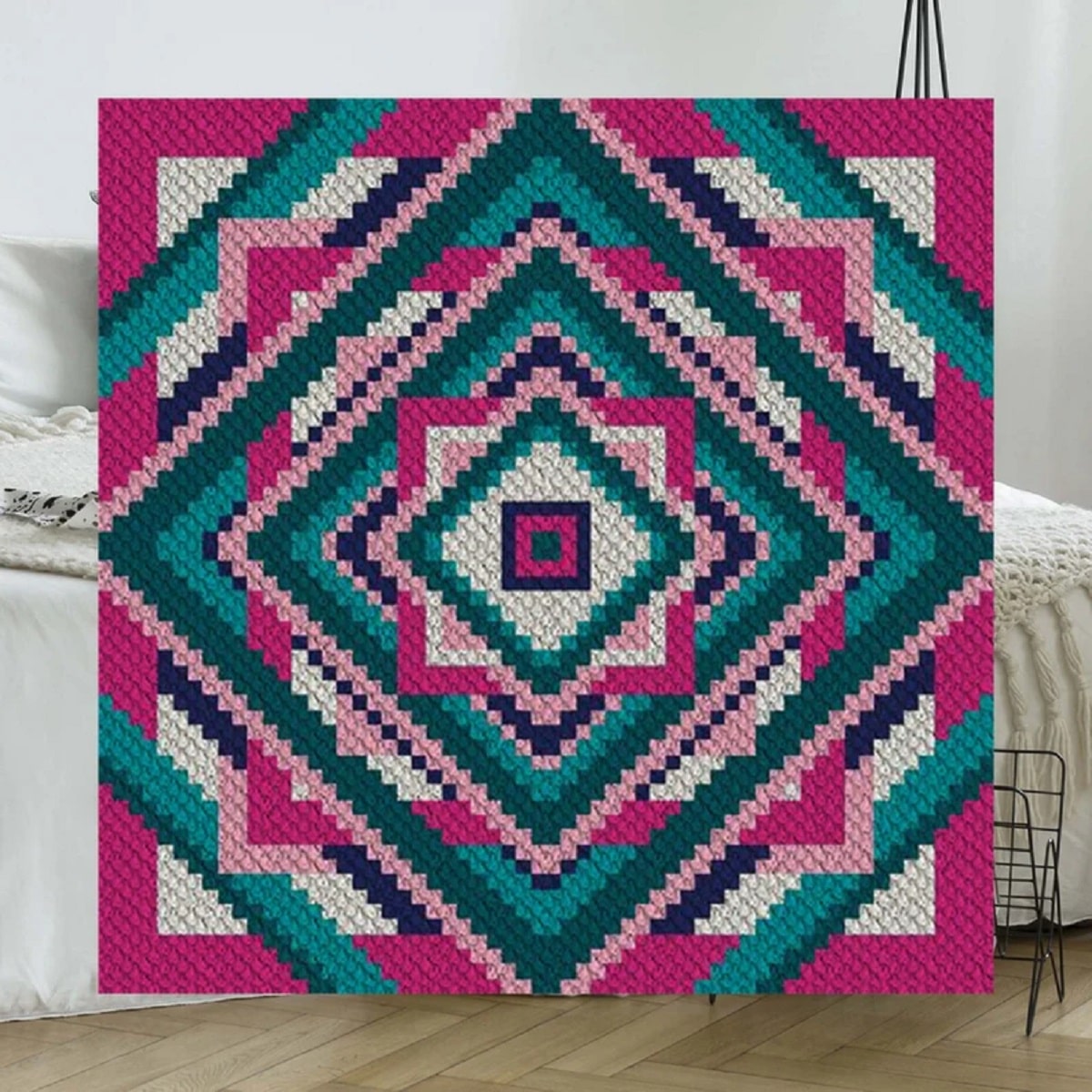 Large crochet afghan square with square and diamond patterns in pink, purple, green, and white in front of a white sofa.