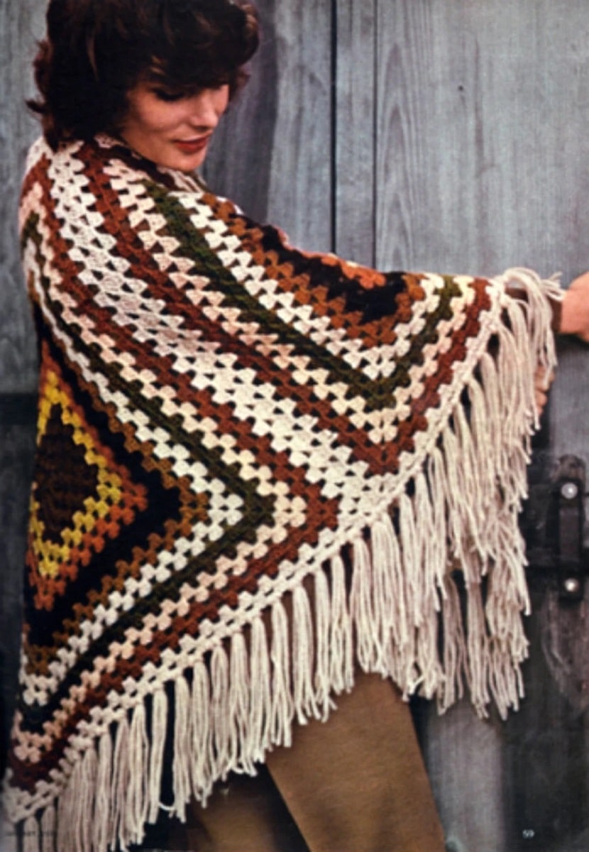 A brunette wearing a white, brown, yellow, and black diamond style shawl with cream fringing in front of gray wood.