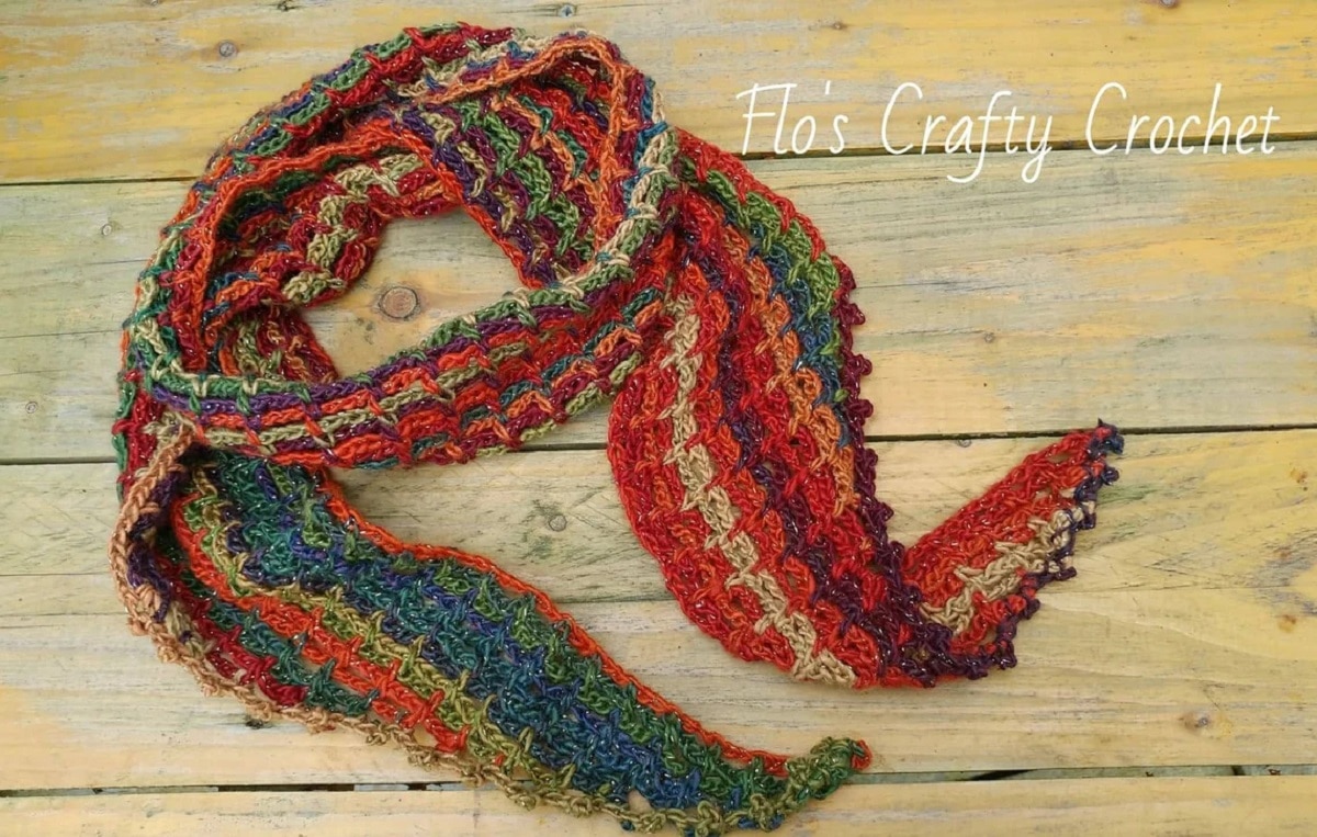 A rainbow colored crochet scarf with vertical stripes twisted into a circle on some pale wooden flooring.