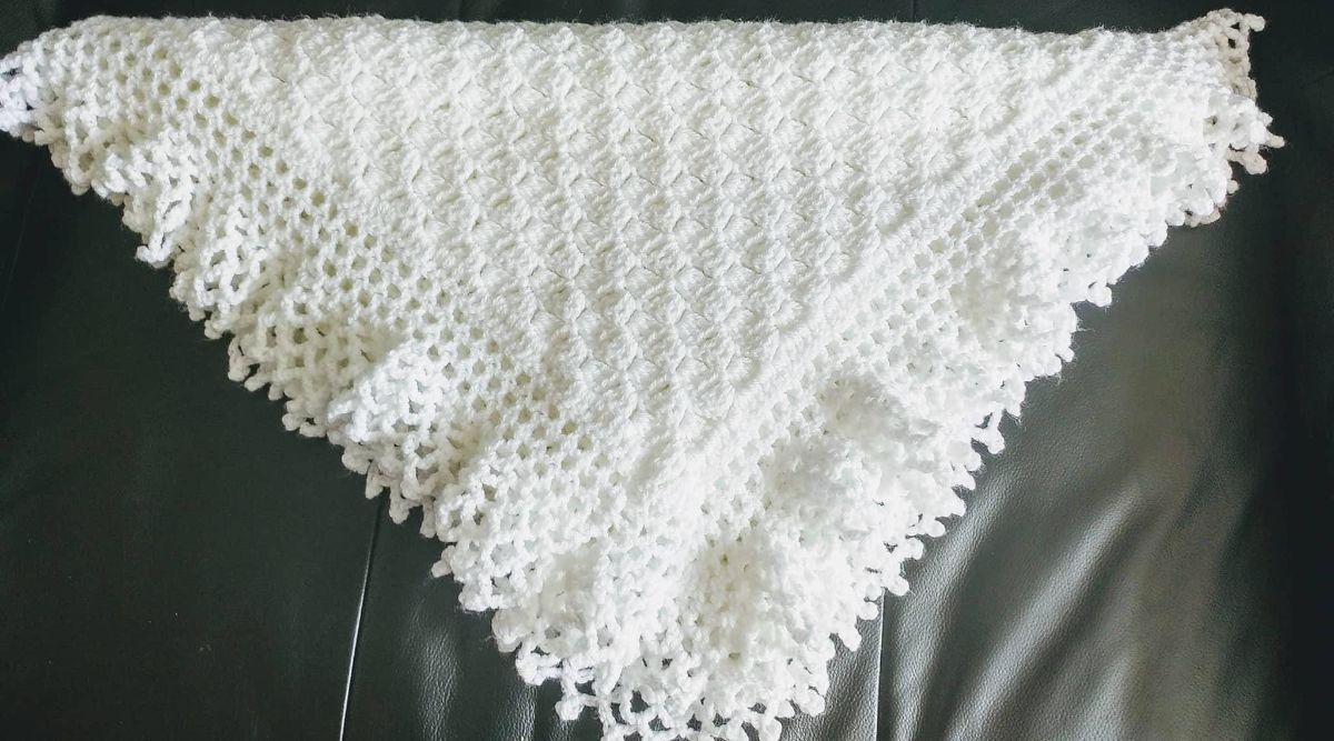 A white crochet blanket with a lacy trim on all sides folded into a triangle on a black couch.