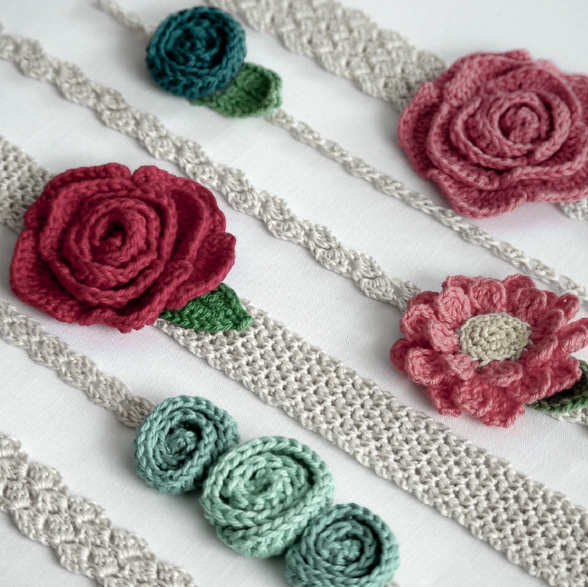 White crochet headbands with a braided design with pink roses, flowers, and small blue knots on them laid across a white background.