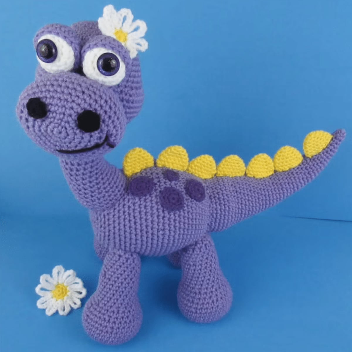 Crochet purple stuffed dinosaur with large black and white eyes, yellow spikes down its back and tail, and a daisy on its head.