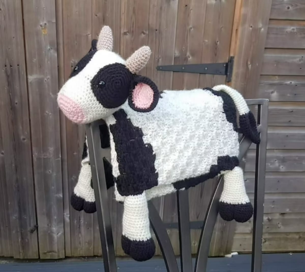 White and vlack crochet cow blanket with a cow’s head and legs attached draped over a brown wooden chair.