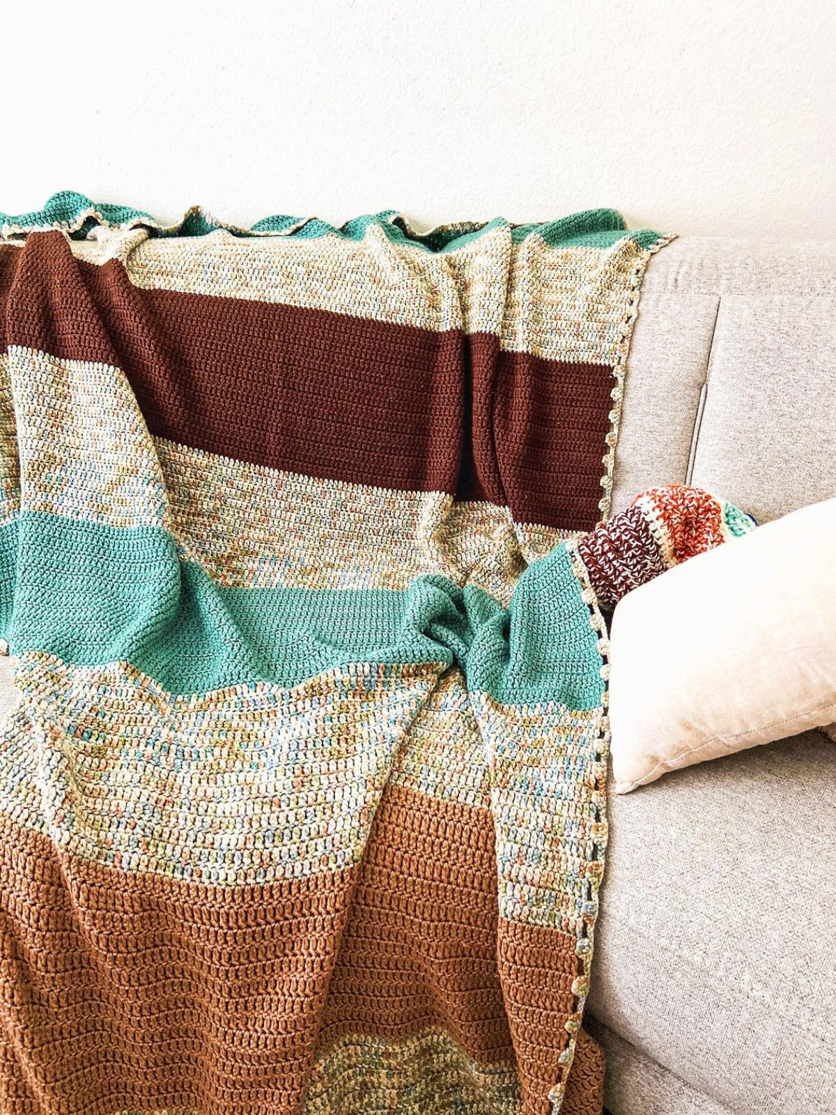 A brown, cream, orange, and blue horizontal striped crochet blanket draped over a cream sofa next to a white cushion.
