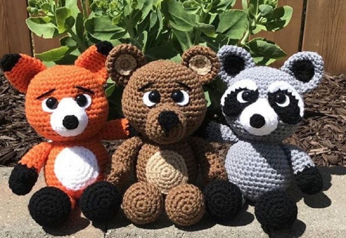 An orange crochet stuffed fox sat next to a brown bear and a gray and black raccoon by a green shrub.