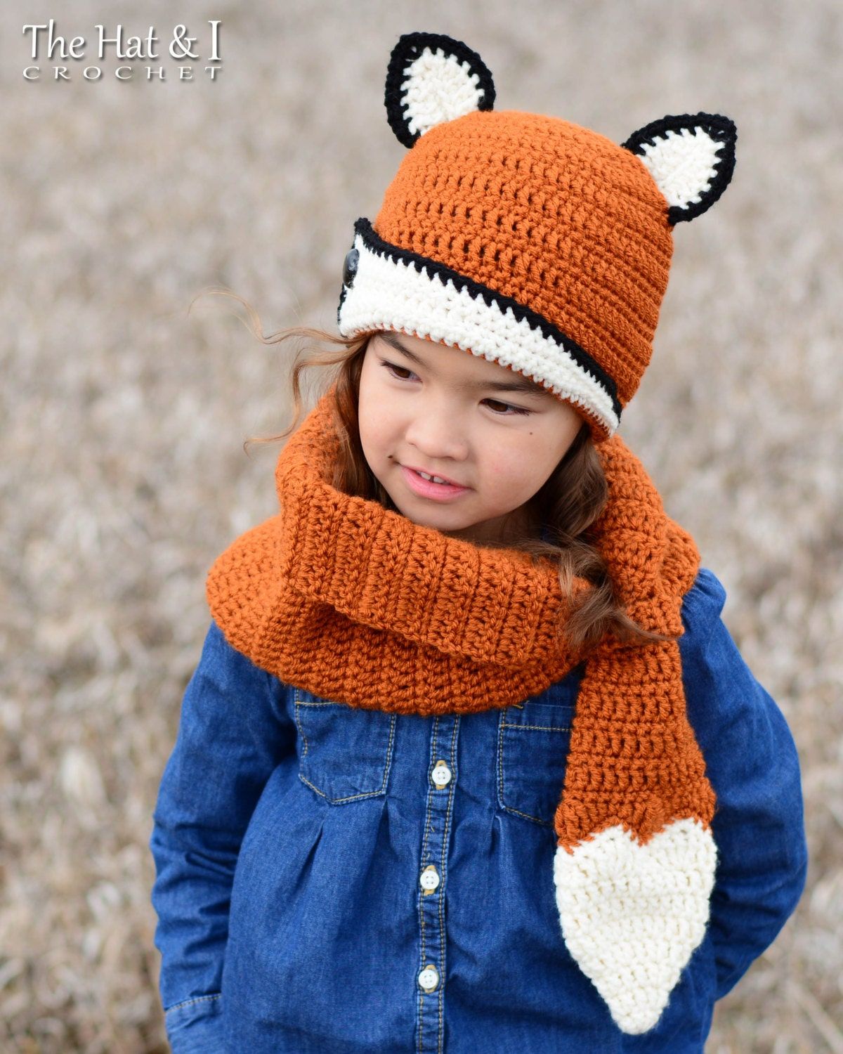 Brunette child outside wearing an orange crochet beanie with white and black fox ears and matching scarf with white fox tail tip.