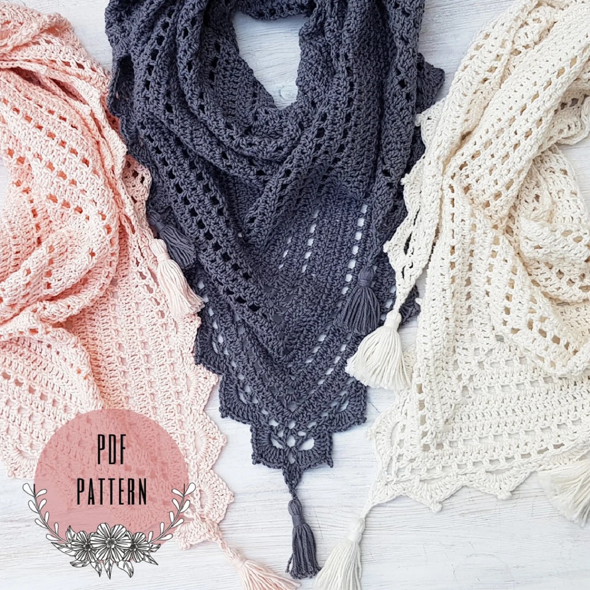 Pale pink, gray, and cream crochet shawls with a zig-zag trim and a tassel at the bottom laying next to each other.