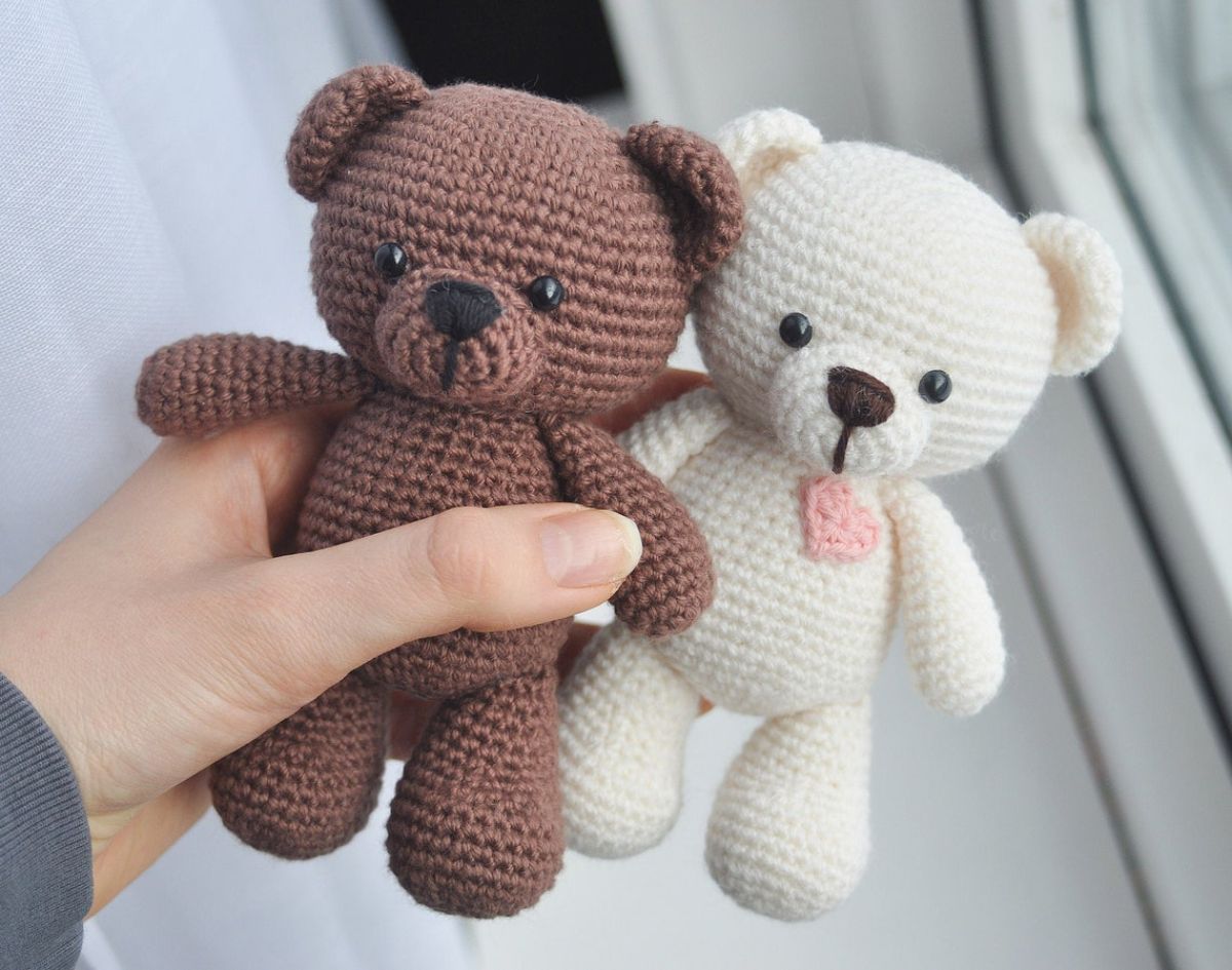 A hand holding a small dark brown crochet teddy bear and a white teddy bear with a small pink heart on its chest.