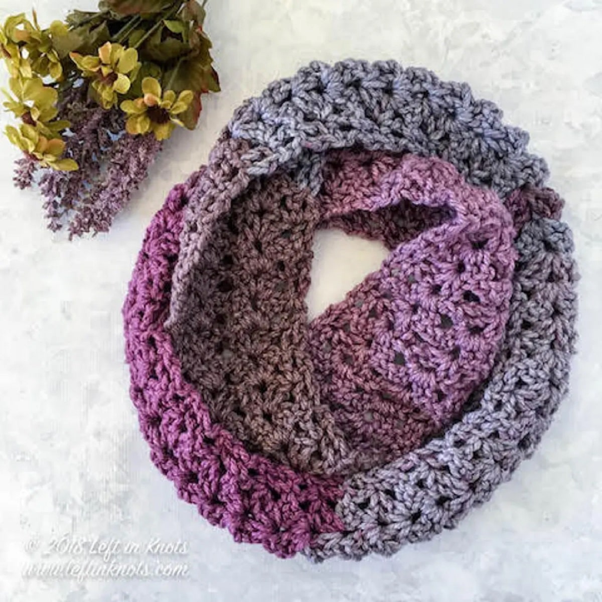 Light purple, dark purple, and gray crochet infinity scarf wrapped into a circle on a white background next to some yellow and purple flowers.