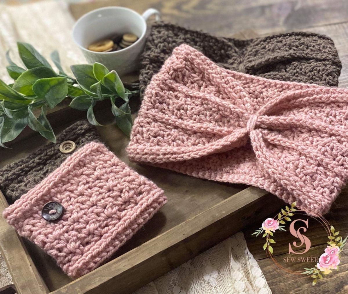 Pale pink ear warmer with folded design and matching scarf on top of a brown warmer with the same design on a brown tray with foliage and a mug nearby.