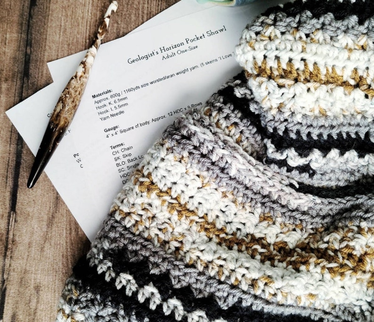 A blue, cream, yellow, and gray striped crochet shawl folded on a wooden table next to some papers and a pen.