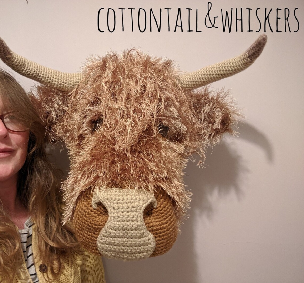 Woman with glasses standing next to a large crochet Highland Cow head mounted on a white wall.