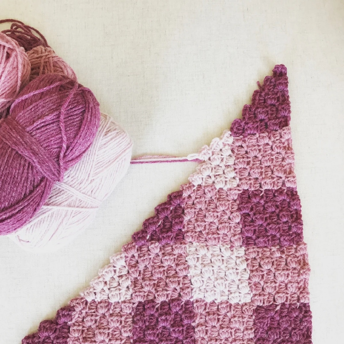 A small triangle of a purple, pink, and white gingham style crochet blanket with balls of yarn in the same color next to it.