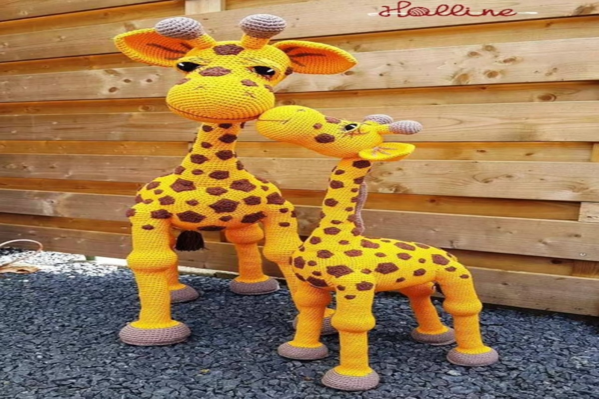  Two bright yellow crochet giraffes with brown spots on their bodies and pale gray hooves on gray flooring by a wooden fence.