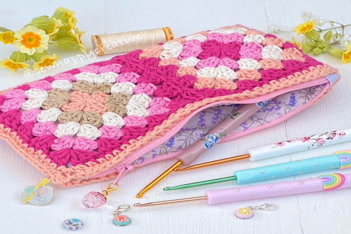 Pink crochet zip up pouch with orange, cream, pale pink, and pink balls stitched onto the top. Zip is open with crochet hooks poking out. 