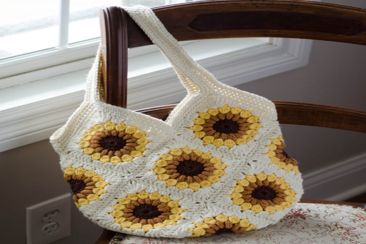 White crochet bag with sunflowers stitched all over it and thick white straps draped over a dark wooden chair.