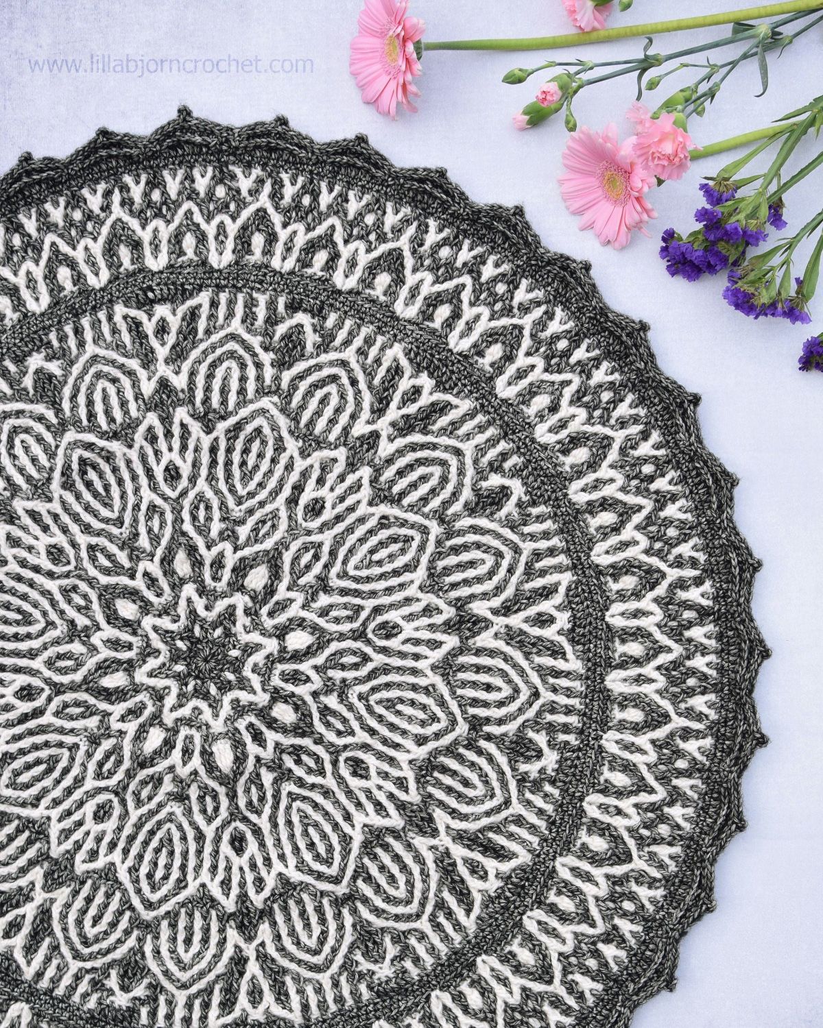 Gray and white round mandala with petal-like designs moving outwards to a narrow gray trim on a white background with pink and purple flowers.