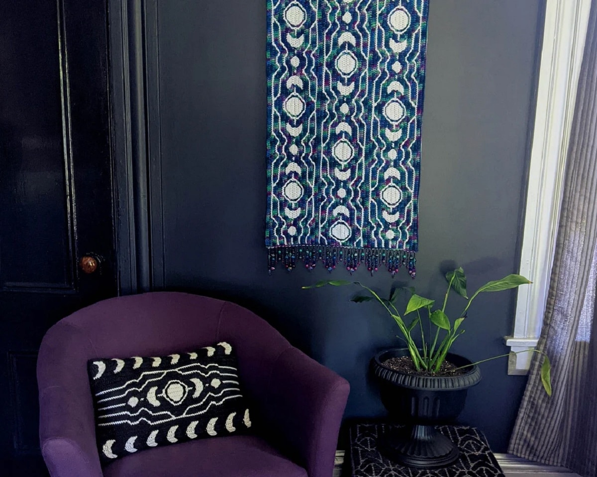  A blue, black, and purple striped crochet wall hanging with white circles running down the rows next to a small black cushion with a similar pattern.