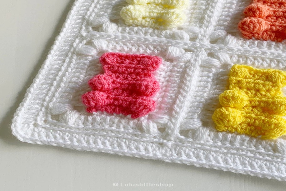 White crochet blanket with pink and yellow gummy bears stitched into the white squares with a white trim around the blanket.