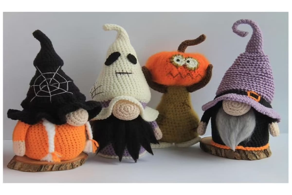 Four crochet gnomes standing in a line, two with witch hats on, one with a ghost hat on and the other a pumpkin on its head.