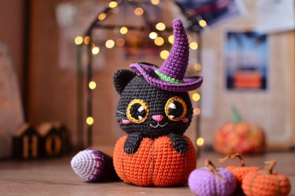 Small crochet stuffed black cat in a purple witches hat and its paws over an orange pumpkin next to smaller pumpkins.