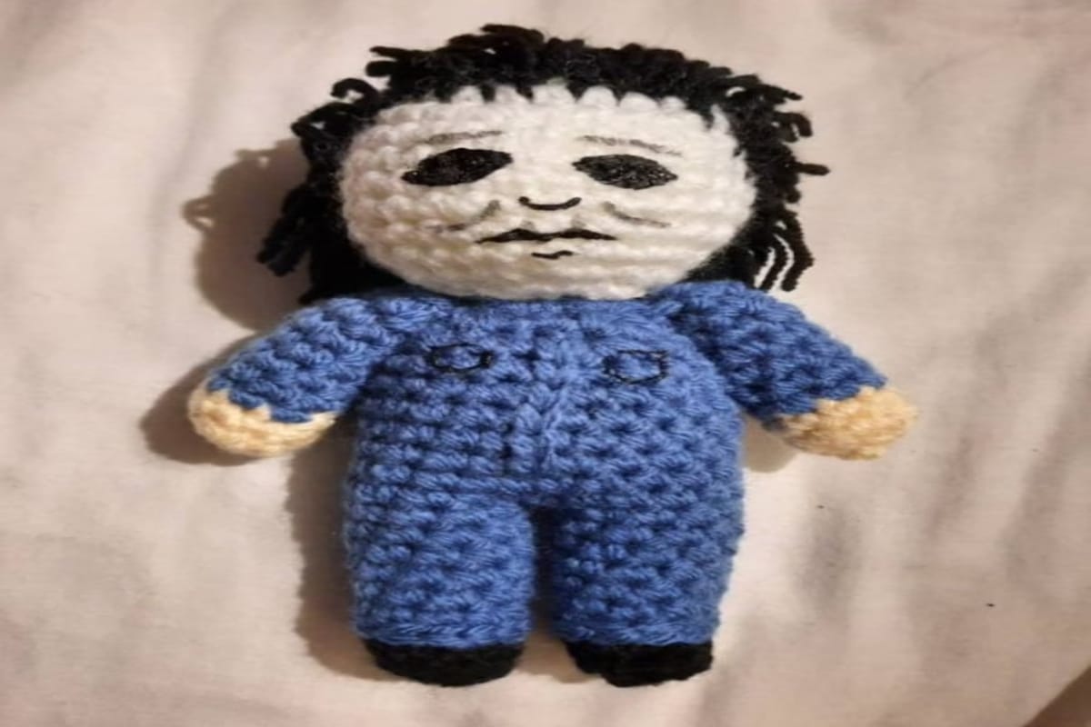 Small crochet stuffed Michael Myers wearing a blue jumpsuit on a white background.