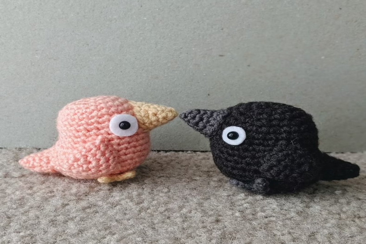 A black crochet crow with a white eye standing beak to beak with a pale pink crochet crow.