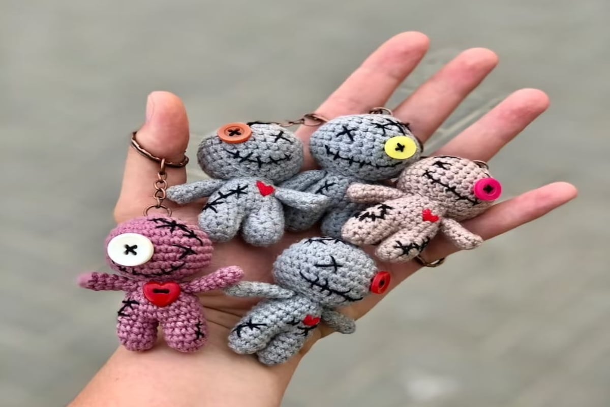 A hand holding purple, gray, and pink miniature crochet voodoo dolls with black stitches on their bodies and faces.