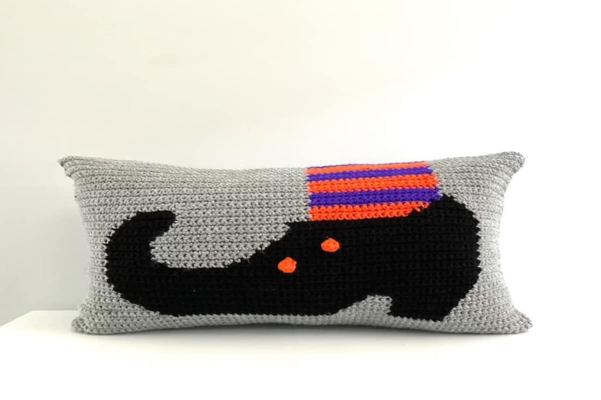 Gray rectangular crochet cushion with a black curled witches shoe and purple and orange striped sock on the front.