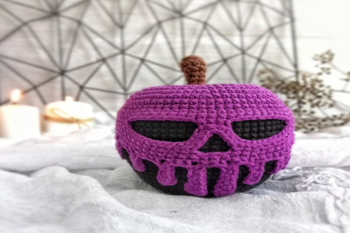 Black crochet apple with a purple skull over the top and brown stem in the middle on a white blanket.