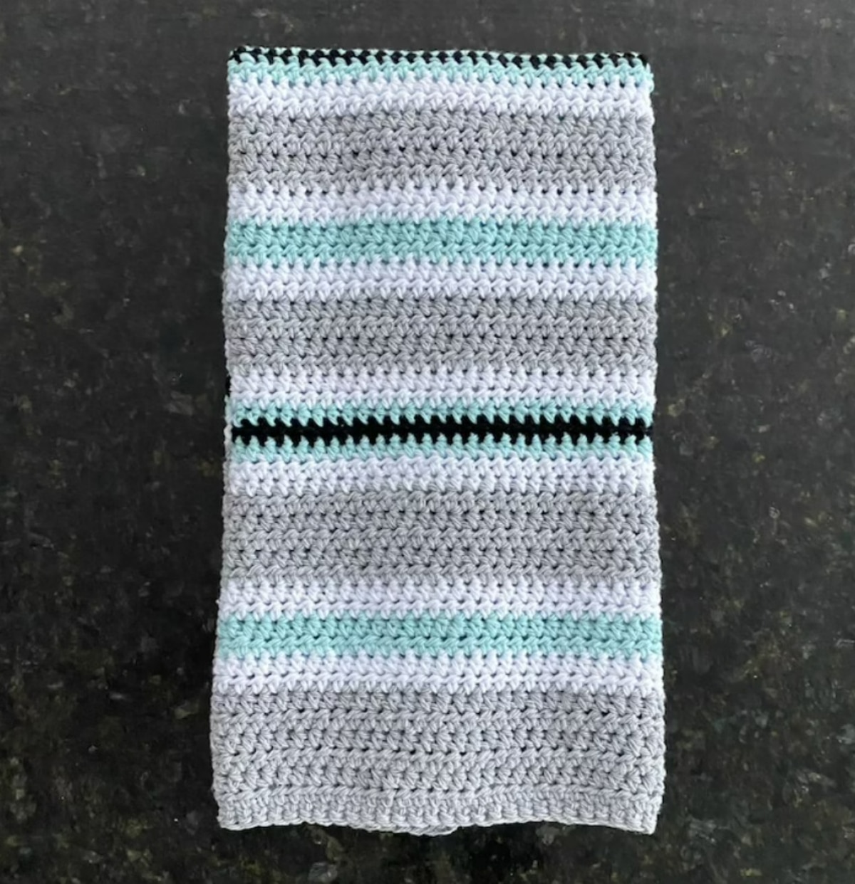 Two square white, gray, and blue horizontal striped crochet dish cloths on a dark gray countertop.