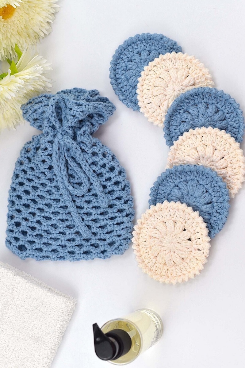 White and blue round crochet scrubbies with a flower in the center next to a small crochet blue bag.