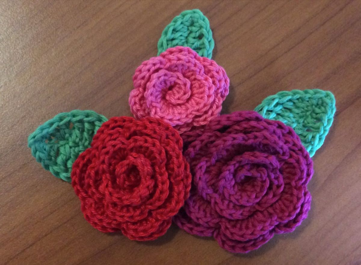 A purple and dark pink crochet rose next to a smaller pink rose with three green leaves on a wooden table.